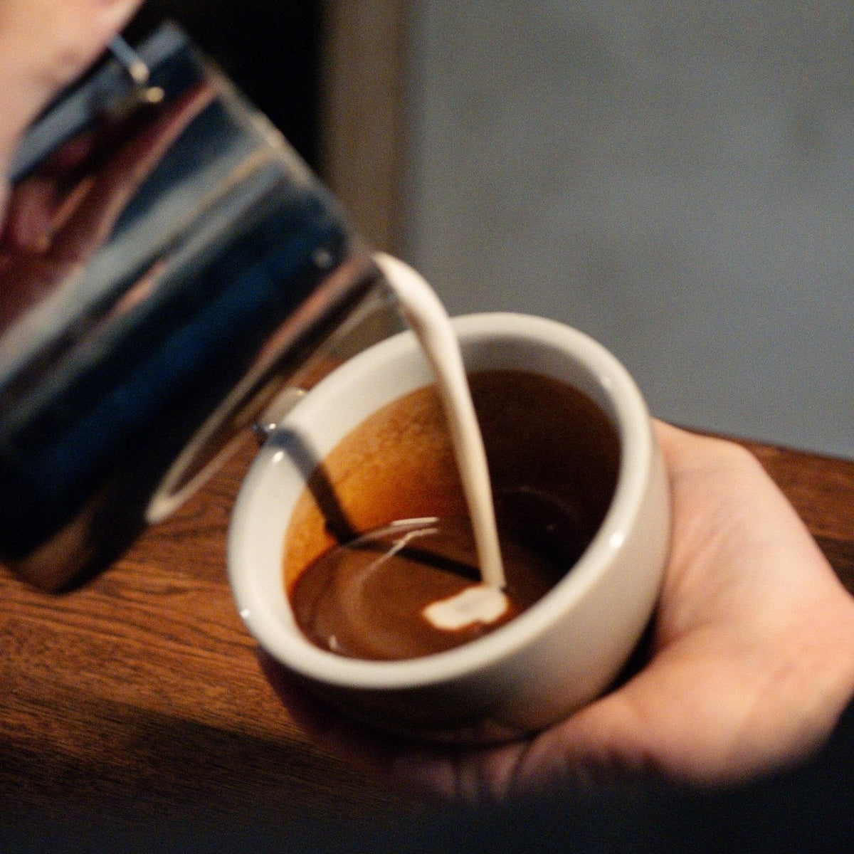 A close-up shot of milk being poured from a metal pitcher into a small white ceramic espresso cup held by a hand, set on a wooden table.