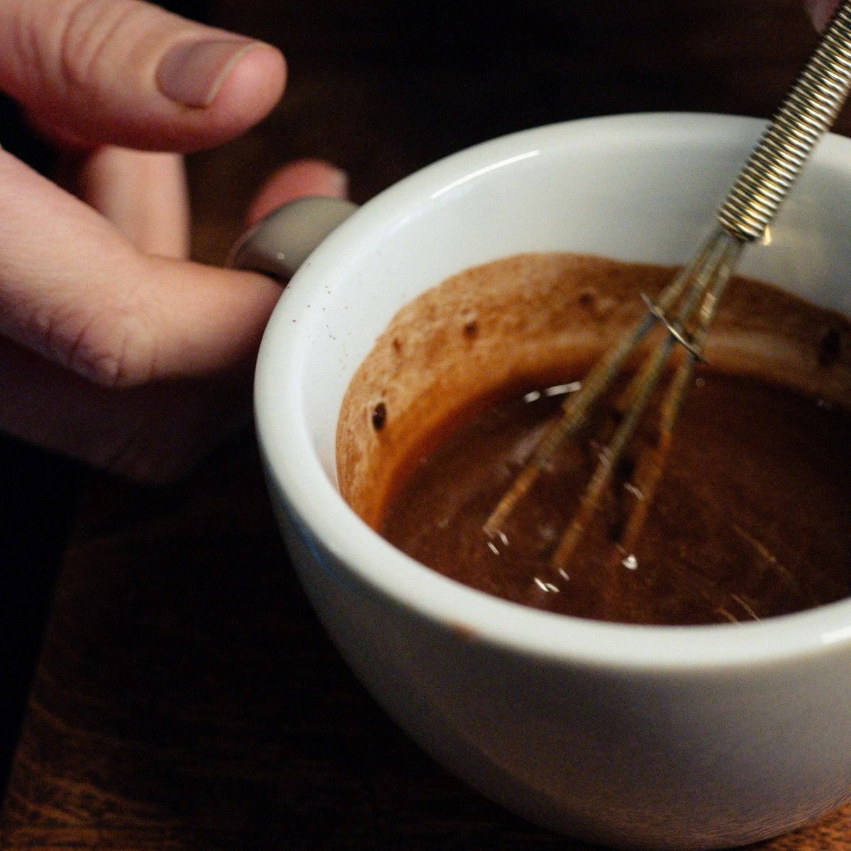 A close-up shot of a white ceramic mug filled with a dark brown chocolate or coffee-based mixture, with a hand holding the handle and a whisk resting inside. The mug sits on a dark, rustic wooden table.