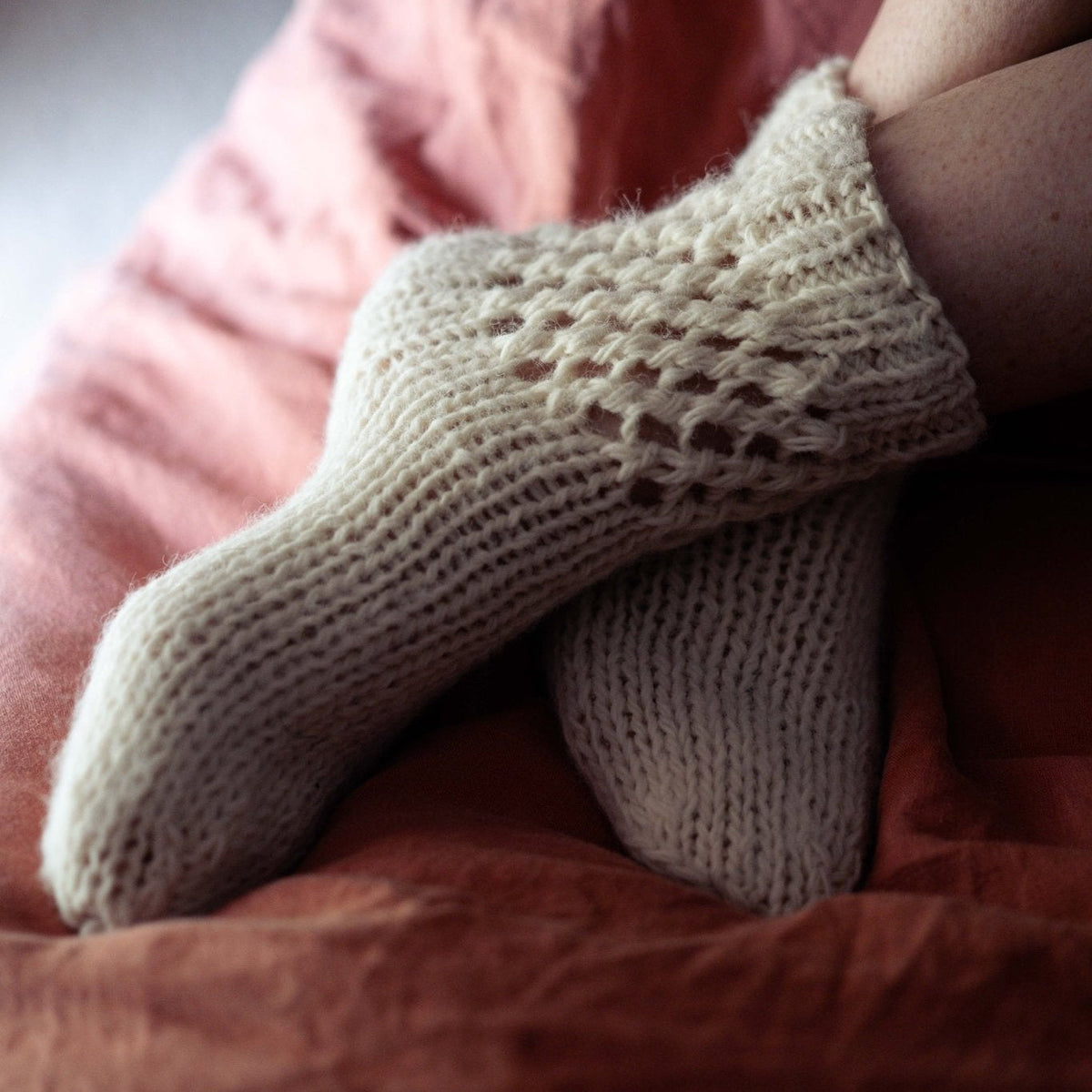 A close-up of a person's feet wearing cream-colored, hand-knit socks while lying on a dusty rose and rust-colored bedspread.