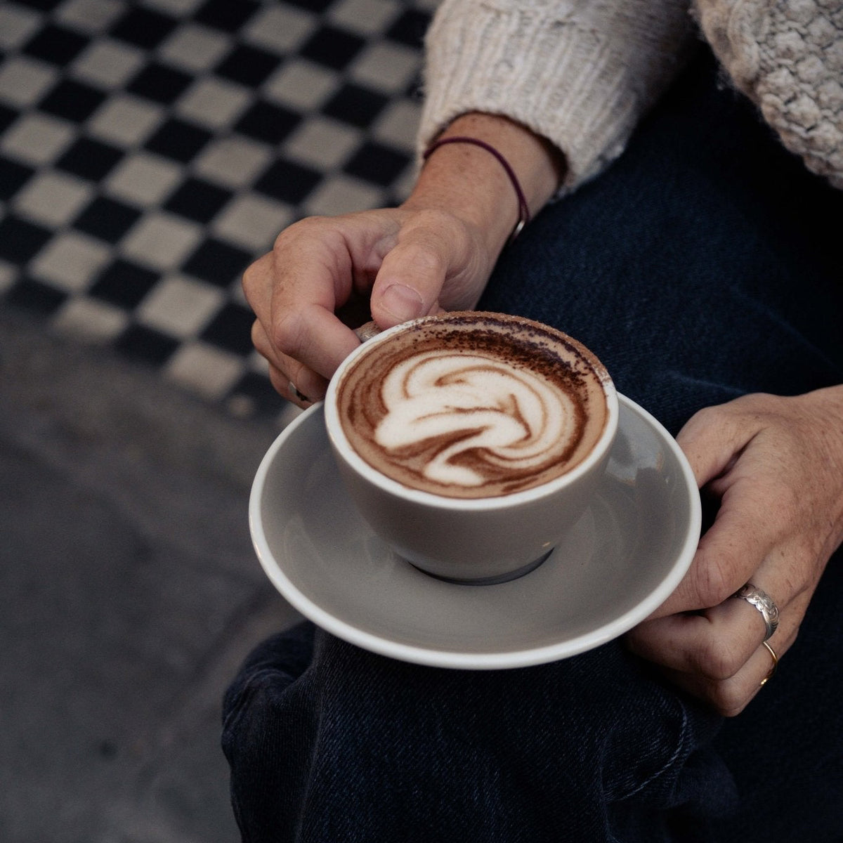 A close-up lifestyle shot of a person holding a white ceramic cup of cappuccino with latte art on a matching saucer, set against their dark jeans and a cream cable-knit sweater.