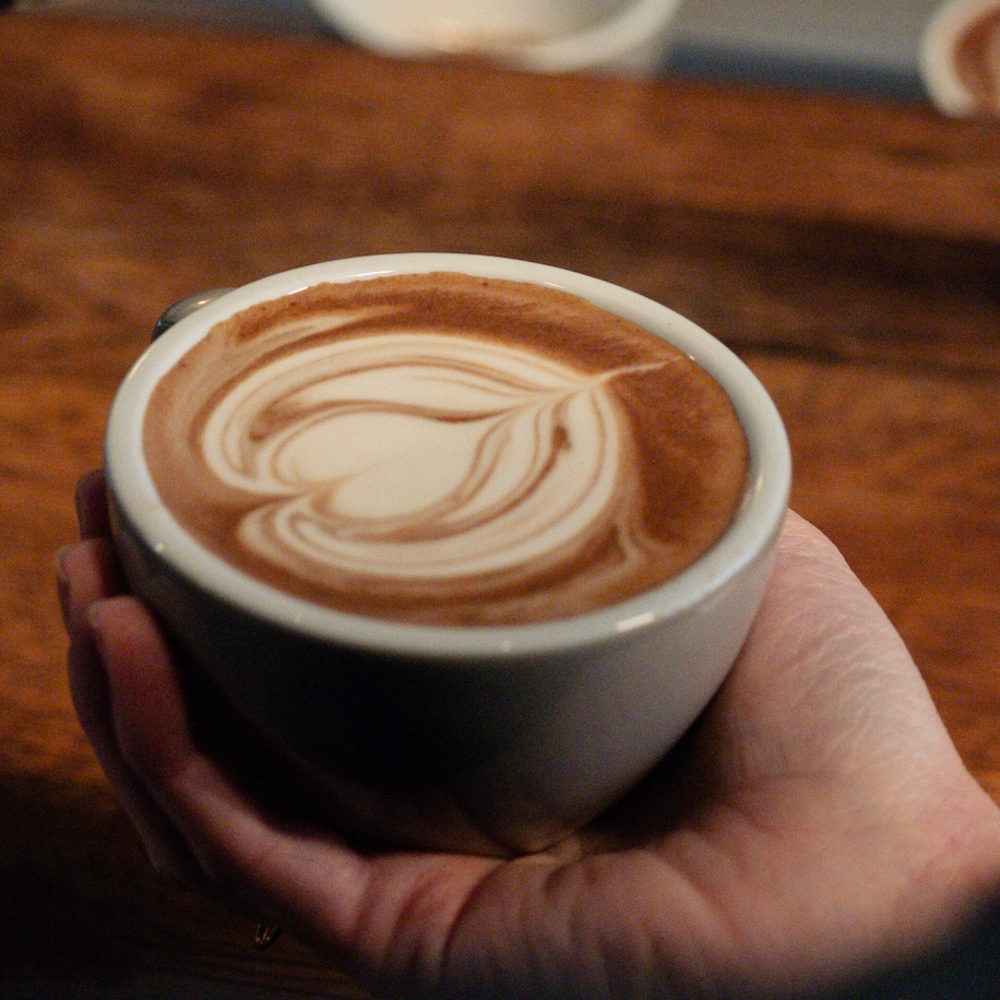 A hand holding a white ceramic cup filled with a hot chocolate featuring a heart-shaped latte art design on the surface, set against a warm wooden table background.