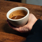 A hand holding a small white ceramic cup filled with a hot chocolate or resting on a wooden table.