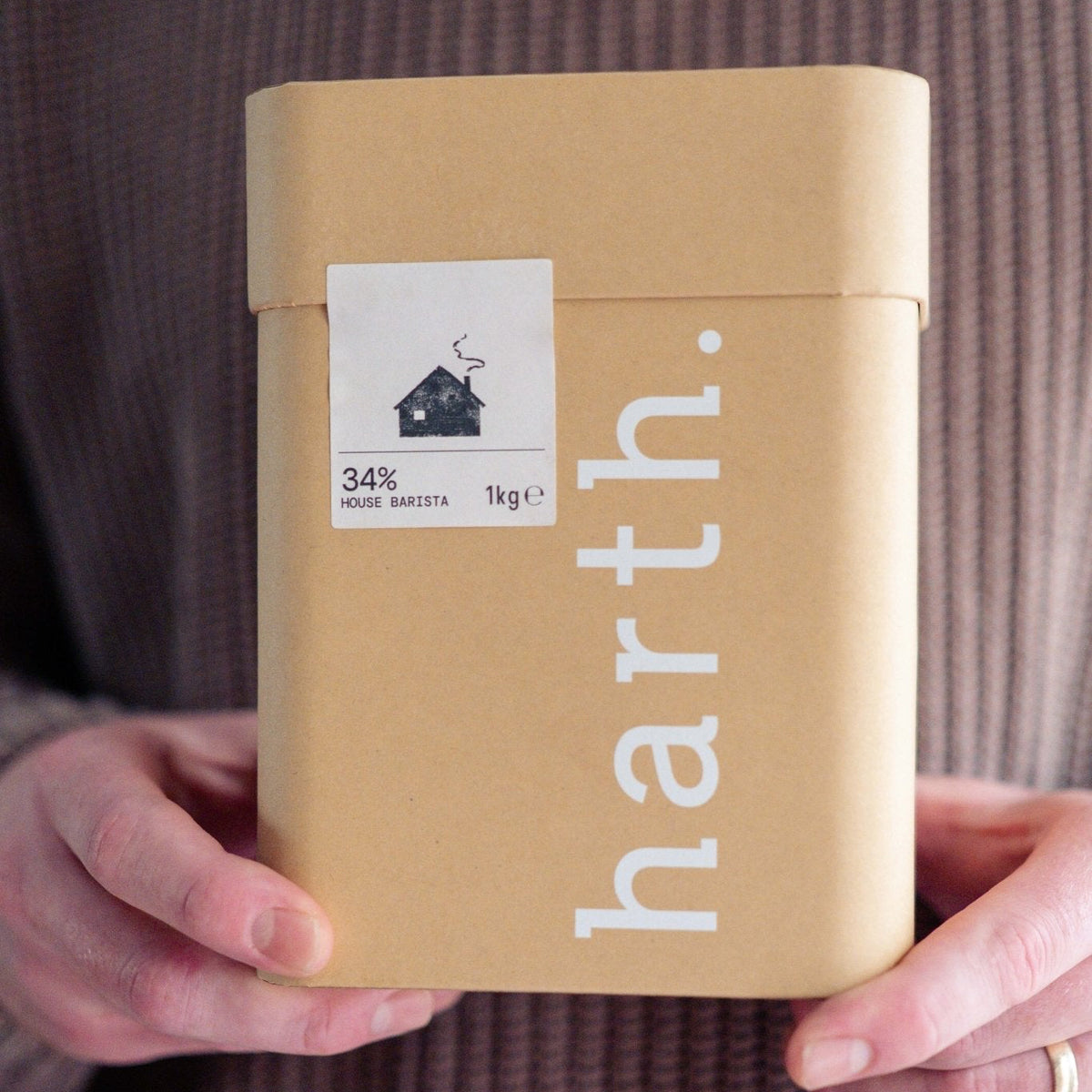 A person in a brown ribbed sweater holds a square kraft paper box with the brand name 'harth.' printed vertically in white.