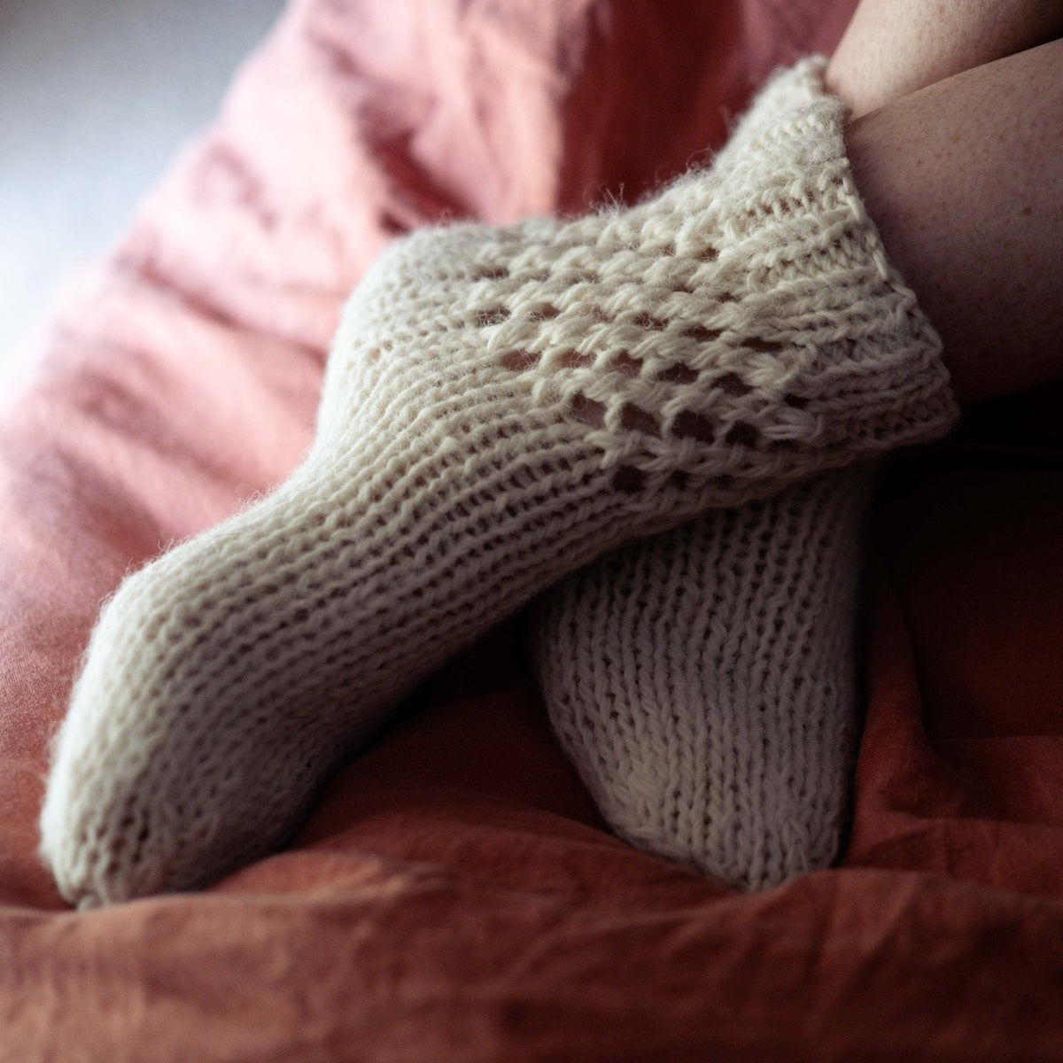 A close-up of a person's feet wearing cream-colored, hand-knit socks while lying on a dusty rose and rust-colored bedspread.